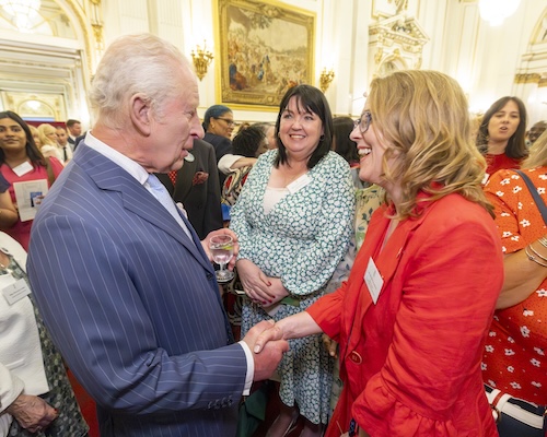 His Majesty The King shaking hands with Ruth Chappell. The King is wearing a blue striped suit, Ruth is wearing a red dress with red jacket